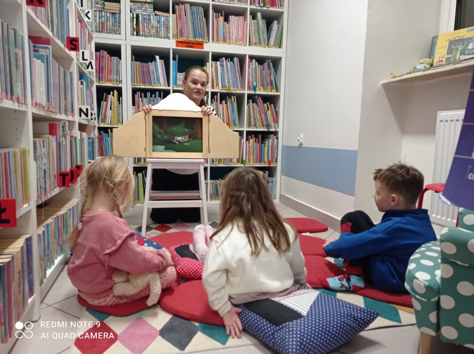 Bibliotekarka otwiera skrzynkę z teatrzykiem kamishibai, widać kilkoro uczestników