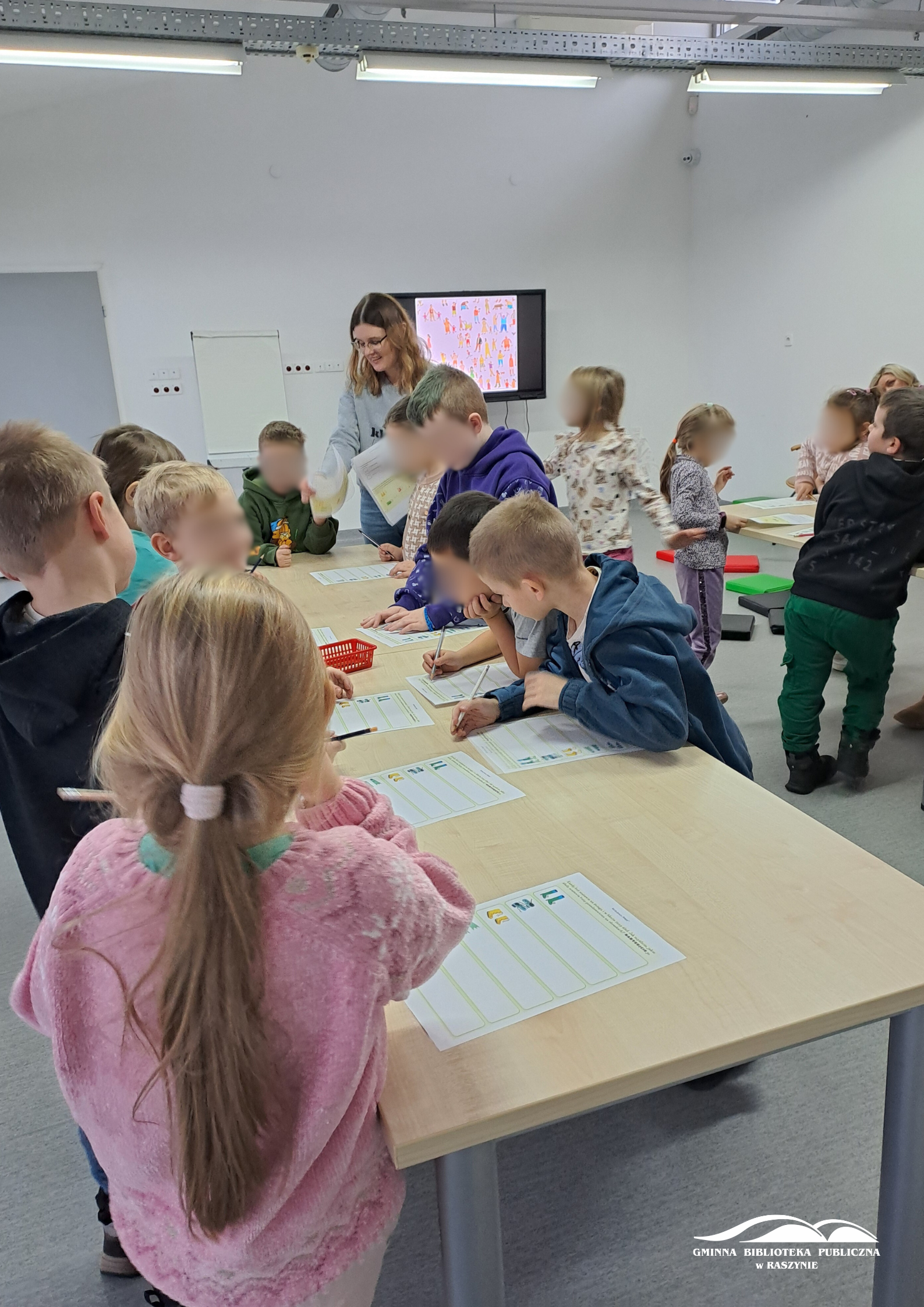 Dzieci stoją przy stolikach i rozwiązują zadania, bibliotekarka rozdaje karty