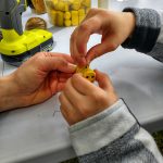 Zbliżenie na dziecięce i dorosłe dłonie tworzące pszczółkę z pomalowanego na żółto korka; w tle widoczne narzędzia, sznurek, pudełko z korkami oraz wkrętarka, sugerujące warsztaty rękodzielnicze.