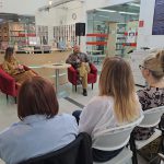 Wojciech Chmielarz i prowadząca siedzą na czerwonych fotelach z mikrofonami w rękach, widać część widowni