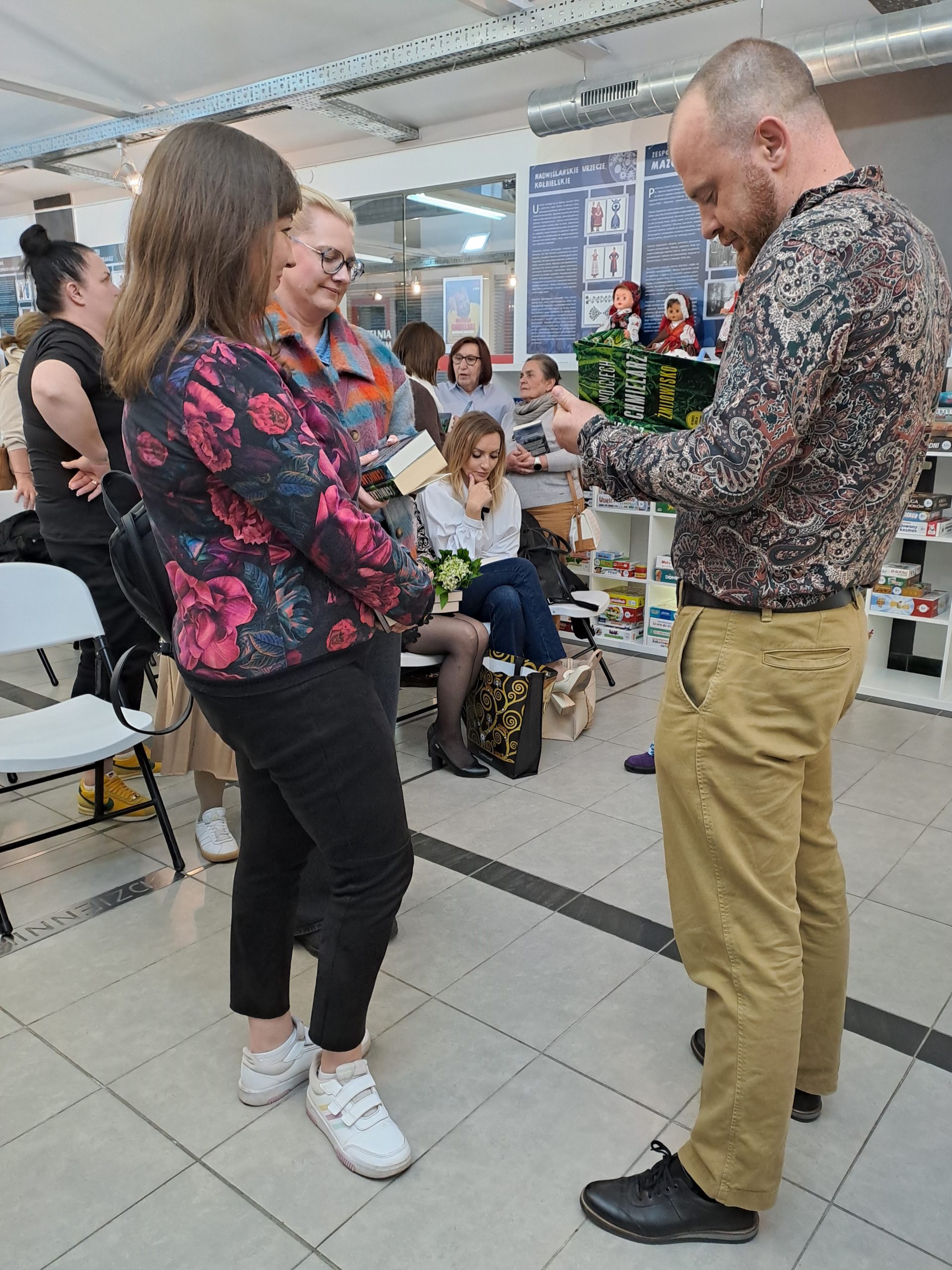Sesja autografów z Wojciechem Chmielarzem - autor podpisuje książki, otoczony przez uczestników spotkania, w tle publiczność