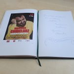 Biblioteczna księga pamiątkowa otwarta na stronie z plakatem spotkania i wpisem: Dziękuję serdecznie za cudowne spotkanie. Pozdrawiam serdecznie Wojciech Chmielarz. Pod wpisem rysunek żmiji.