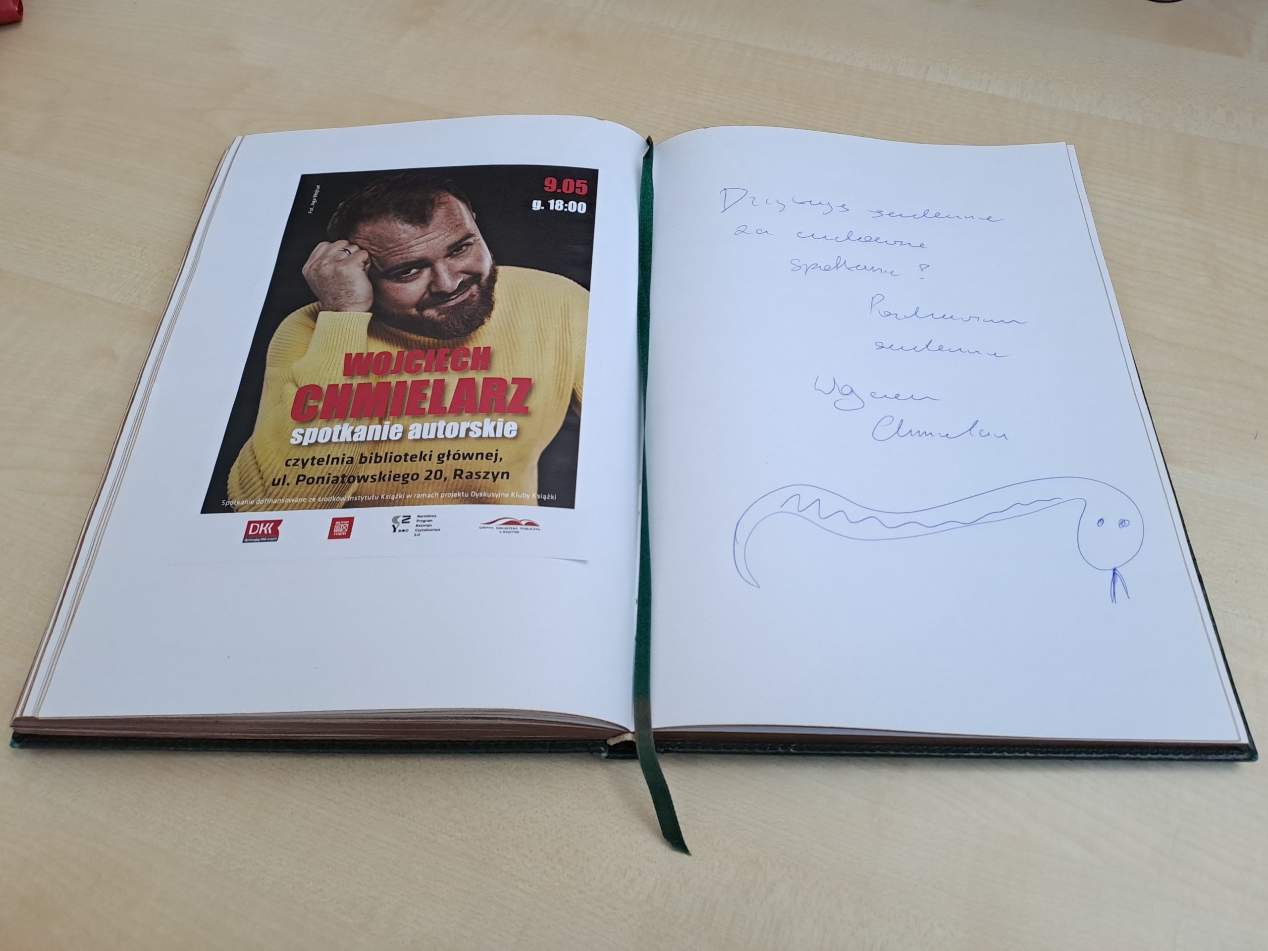 Biblioteczna księga pamiątkowa otwarta na stronie z plakatem spotkania i wpisem: Dziękuję serdecznie za cudowne spotkanie. Pozdrawiam serdecznie Wojciech Chmielarz. Pod wpisem rysunek żmiji.