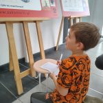 Mały chłopiec siedzi przed planszą wystawy z kartką i długopisem