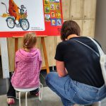 Mama z córką siedzą przed jedną z plansz wystawy, rozwiązują zadania z quizu