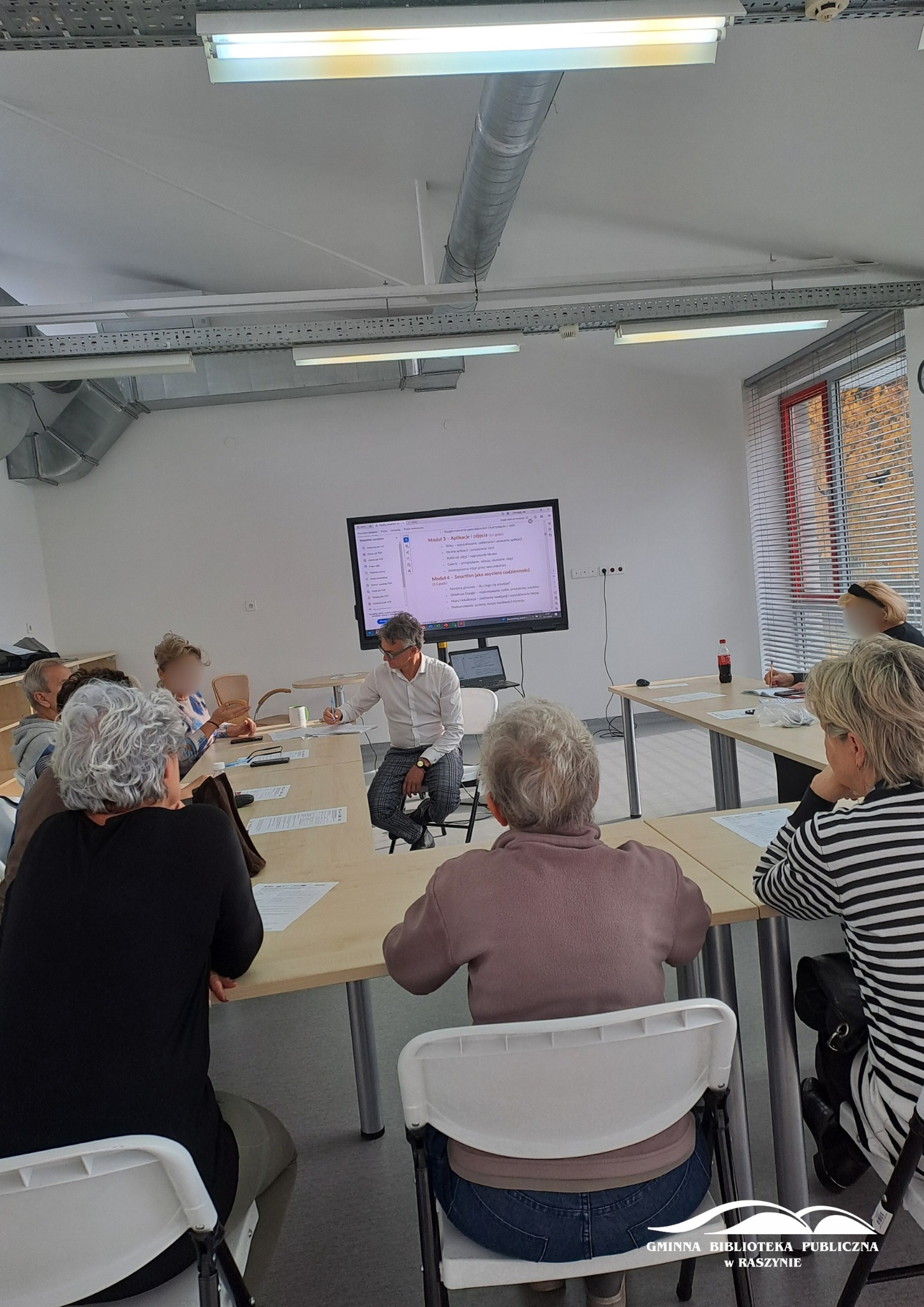 Grupa uczestników zajęć z obsługi smartfona wraz z prowadzącym, który coś tłumaczy