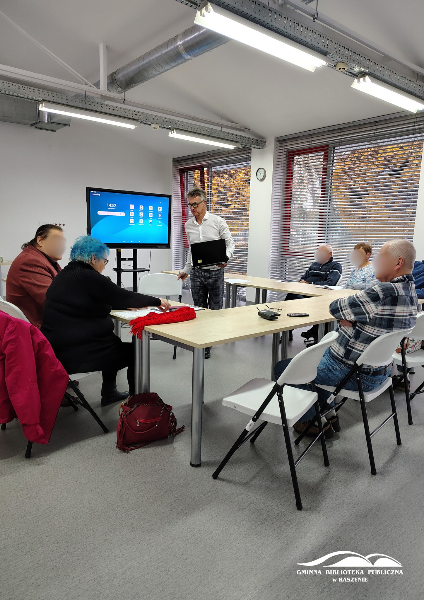 Grupa uczestników zajęć z obsługi smartfona wraz z prowadzącym na tle ekranu multimedialnego