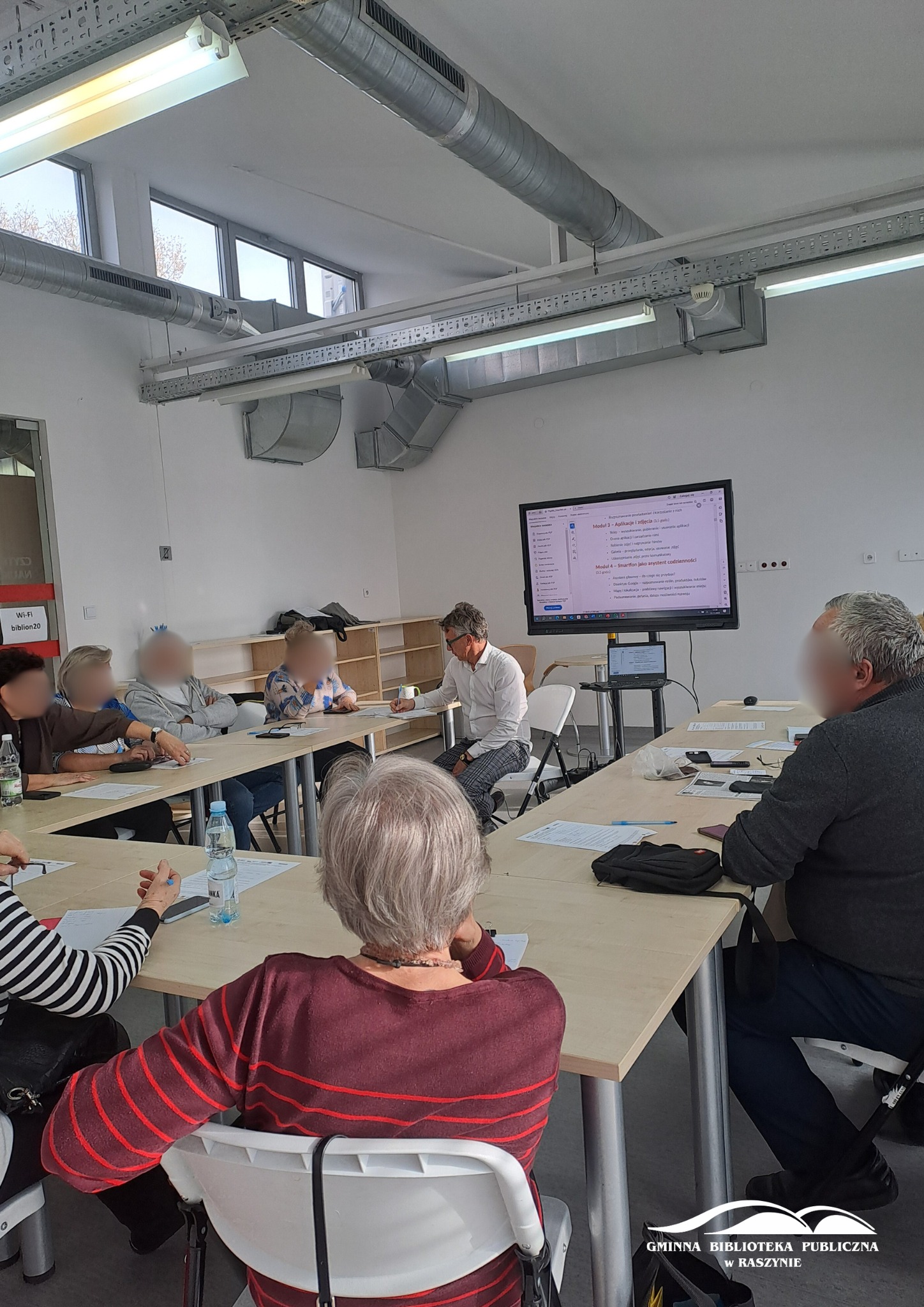 Grupa uczestników zajęć z obsługi smartfona siedzi przy stole wraz z prowadzącycm