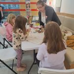 Warsztaty tworzenia kokard patriotycznych - grupa uczestników przy stole, prowadząca tłumaczy