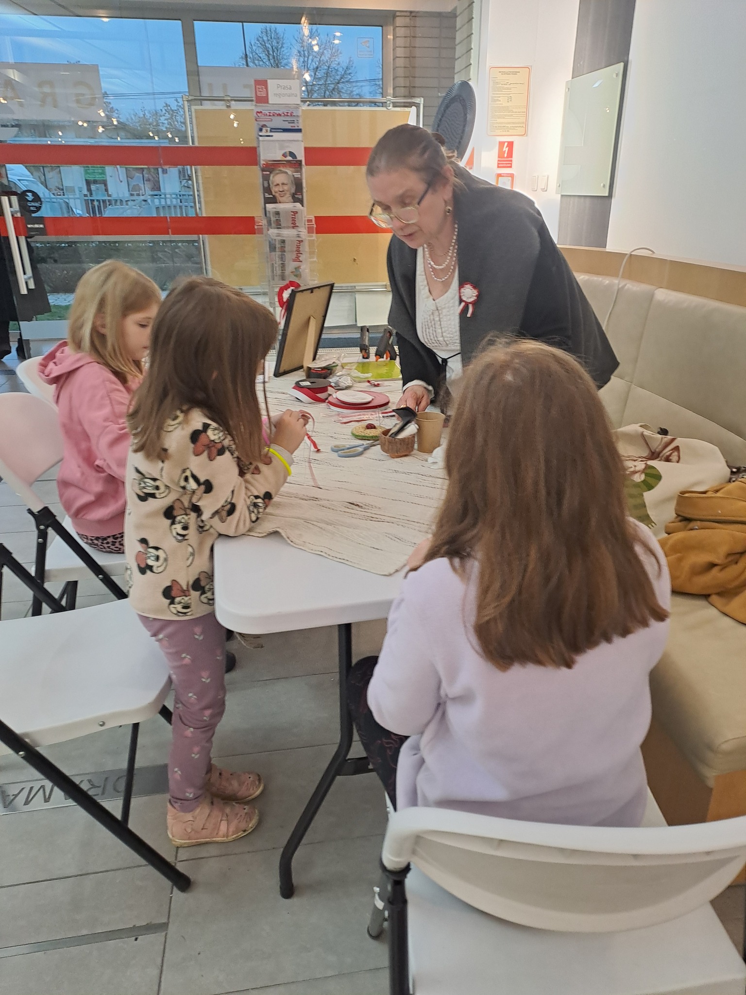 Warsztaty tworzenia kokard patriotycznych - grupa uczestników przy stole, prowadząca tłumaczy