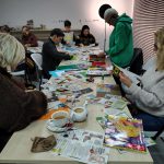 Grupa uczestników warsztatów kolażowych siedzi przy stołach podczas pracy