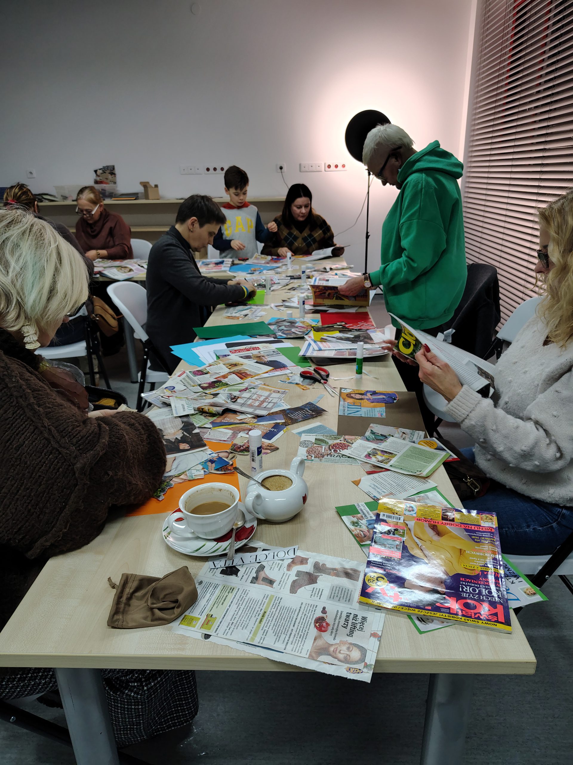 Grupa uczestników warsztatów kolażowych siedzi przy stołach podczas pracy