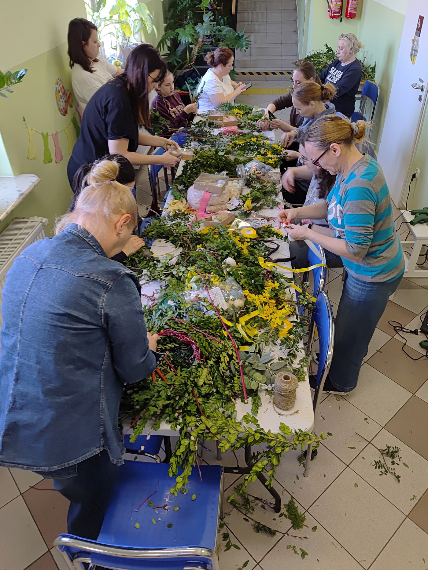 Uczestnicy warsztatów stoją wokół stołów z materiałami do wieńców