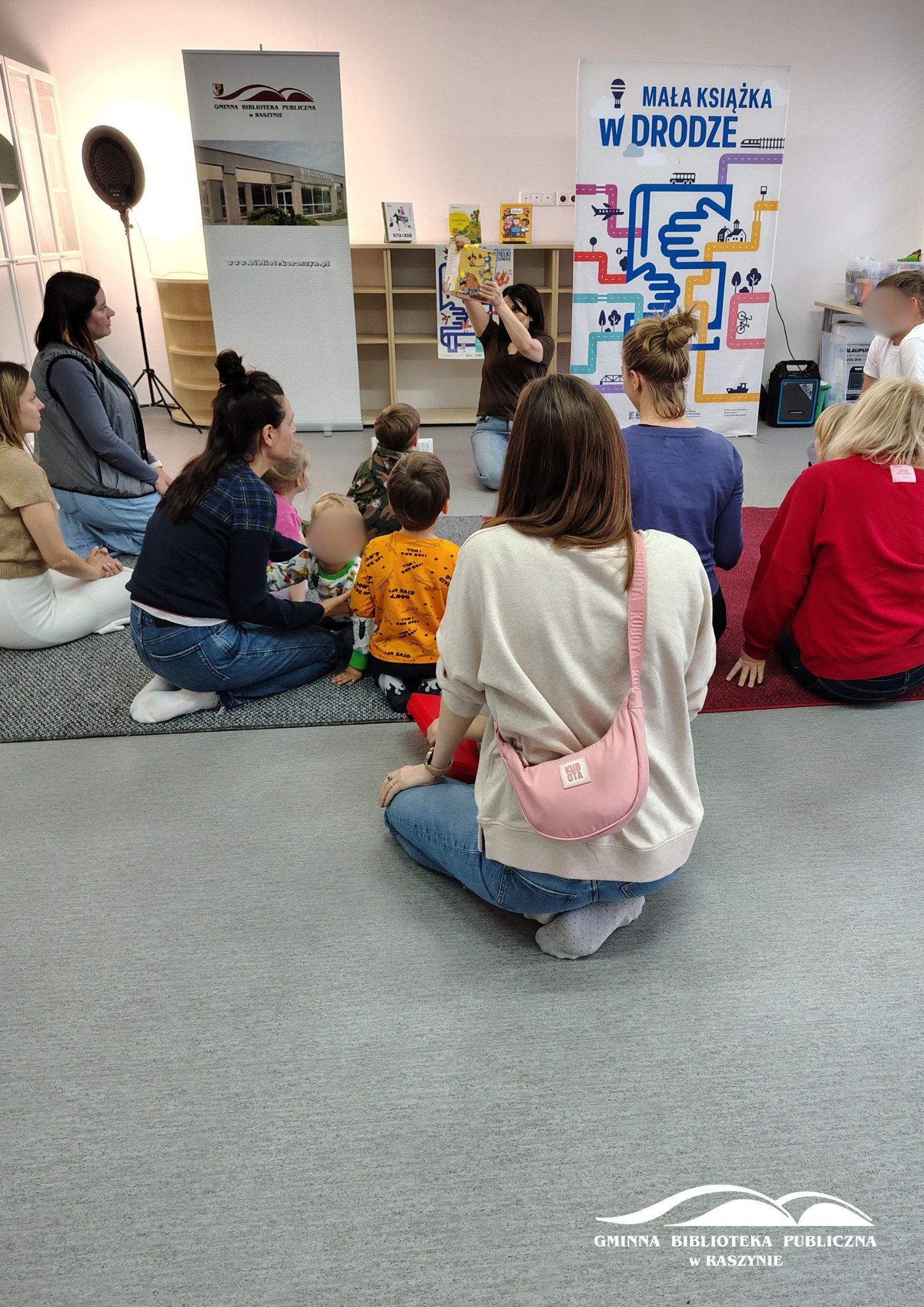 Grupa rodziców z dziećmi podczas warsztatów czytelniczych, prowadząca pokazuje ilustracje w książce