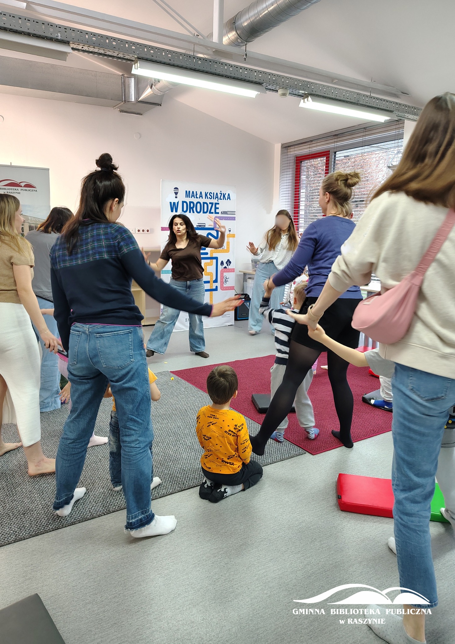 Grupa rodziców z dziećmi podczas radosnego tańca, widoczna też prowadząca zajęcia