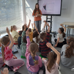 Bibliotekarka stoi przy ekranie multimedialnym, dzieci zgłaszają się z uniesionymi dłońmi do odpowiedzi