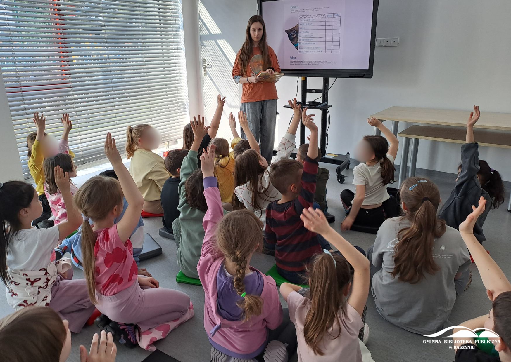 Bibliotekarka stoi przy ekranie multimedialnym, dzieci zgłaszają się z uniesionymi dłońmi do odpowiedzi