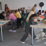 Uczniowie stoją wokół dwóch stołów, rysując. Widoczna prowadząca zajęcia i nauczycielka