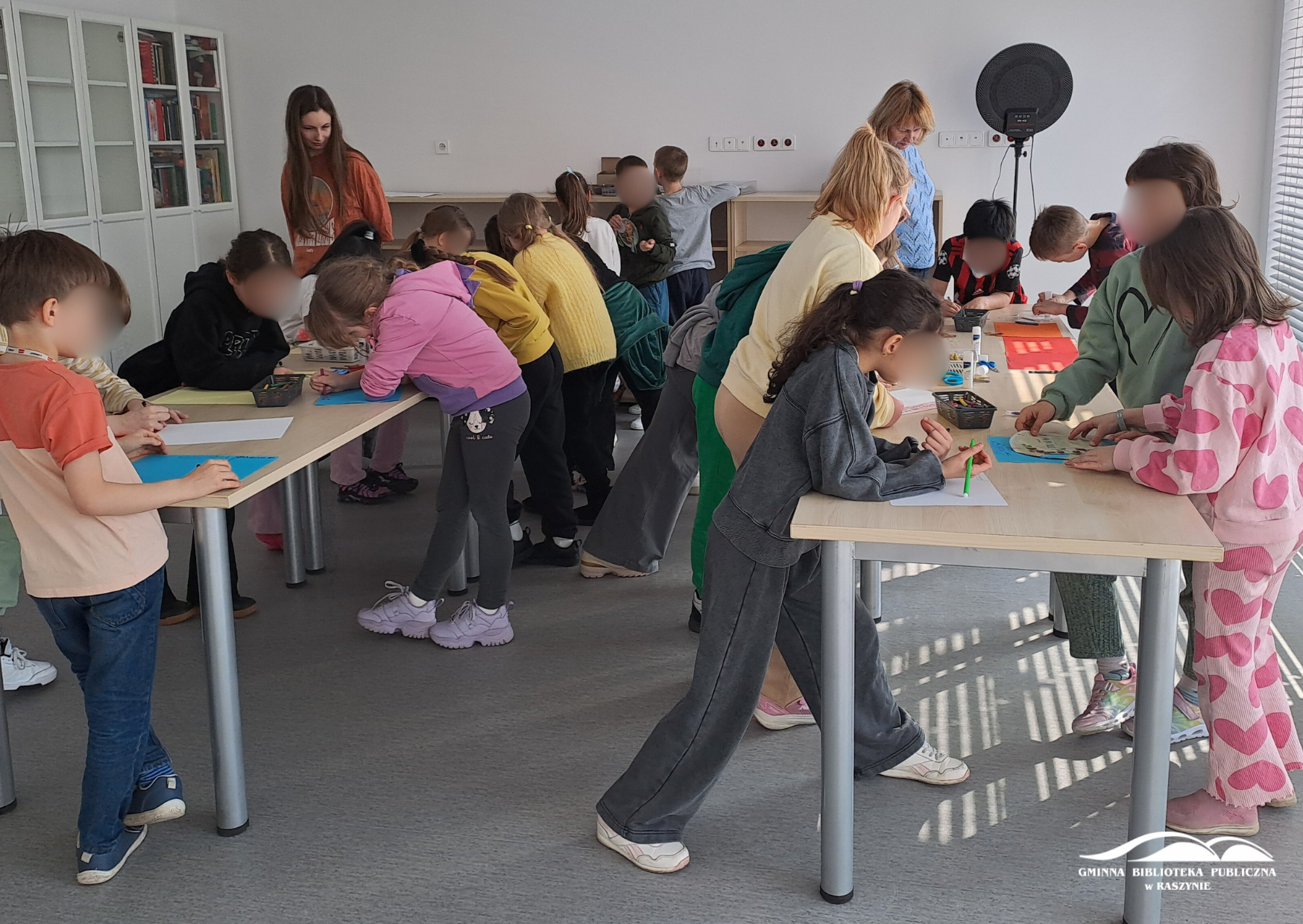 Uczniowie stoją wokół dwóch stołów, rysując. Widoczna prowadząca zajęcia i nauczycielka
