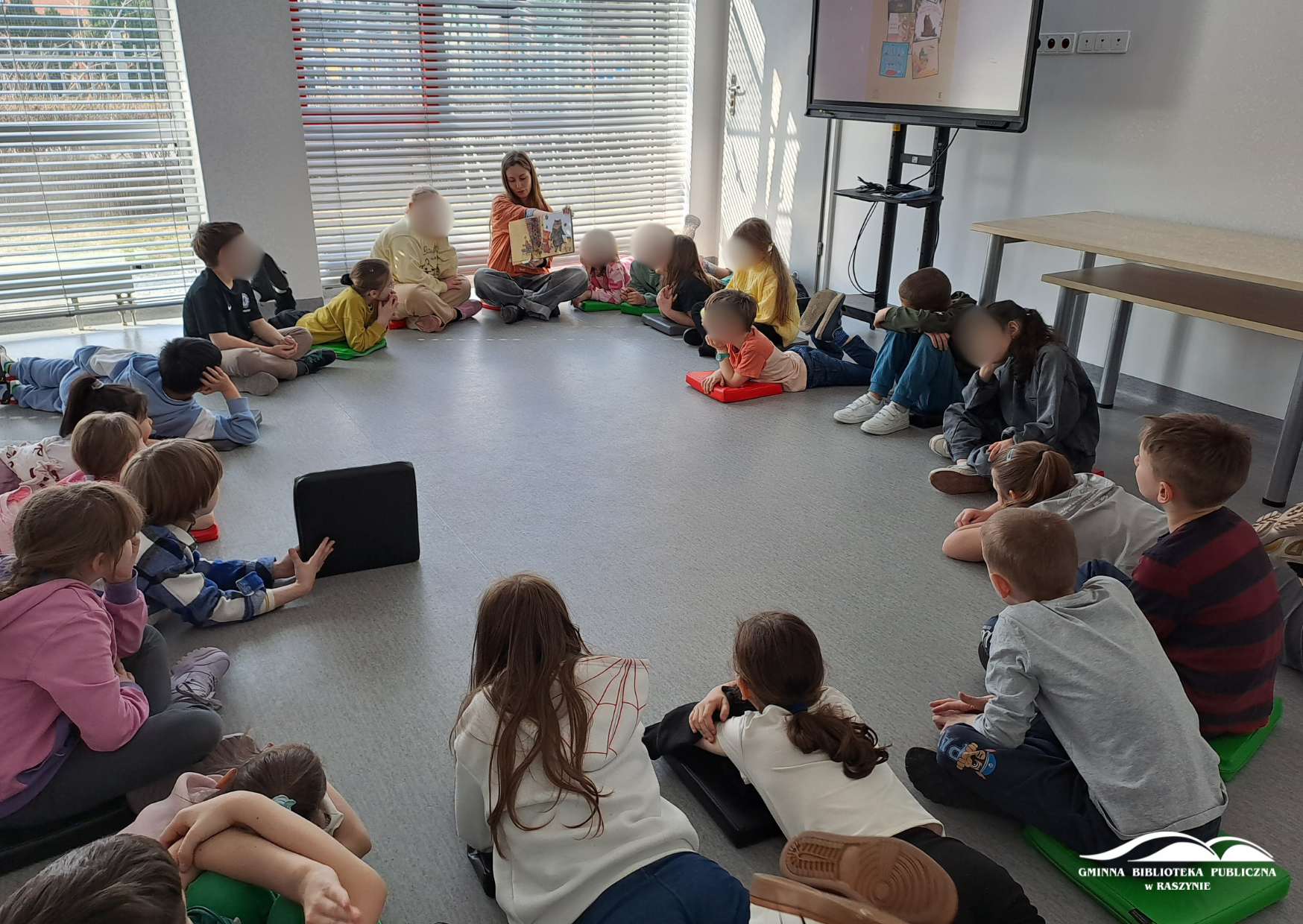 Dzieci siedzą w kole na podłodze, bibliotekarka czyta książkę