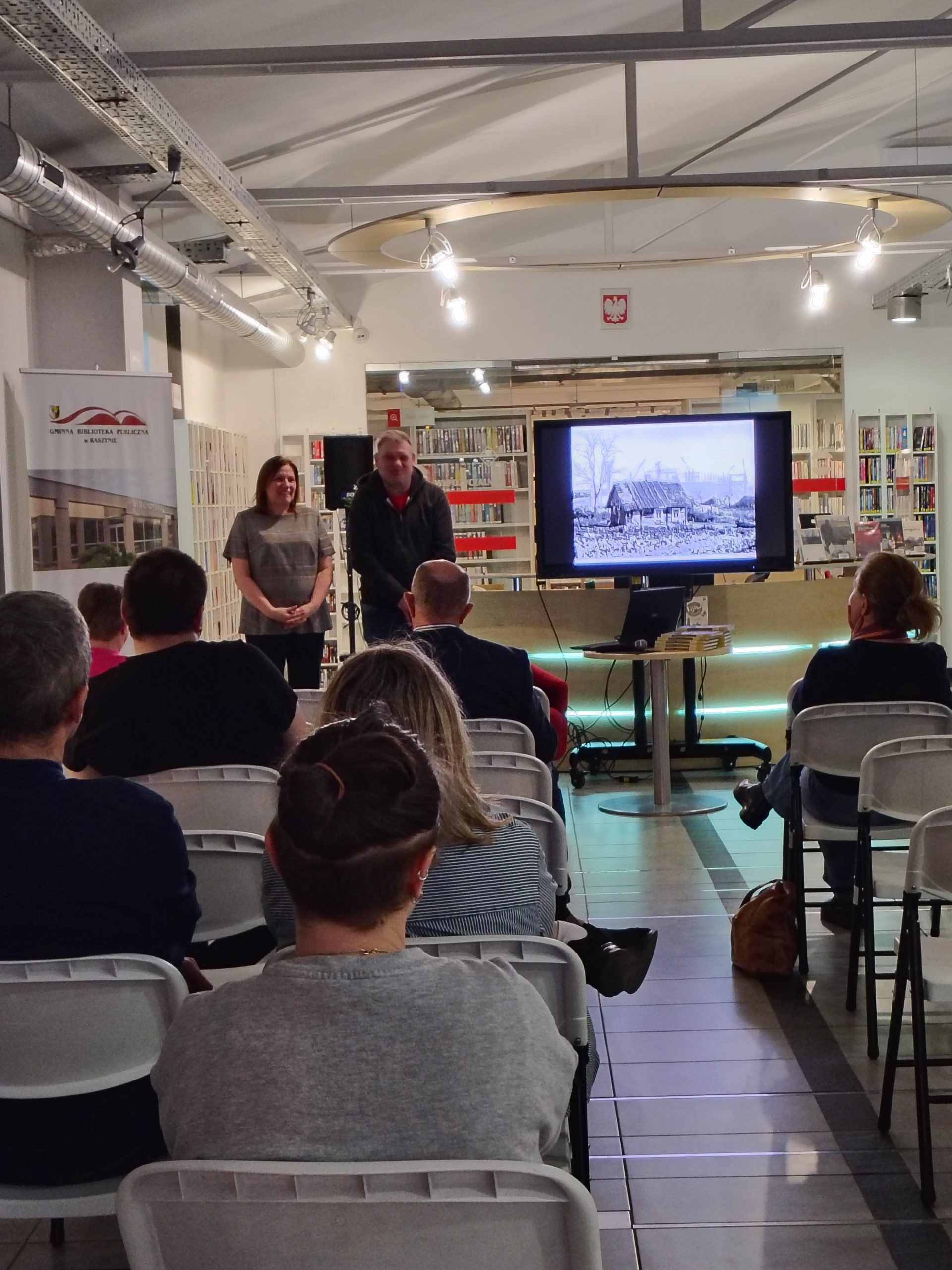 Dyrektor biblioteki przedstawia gościa, widoczna publiczność