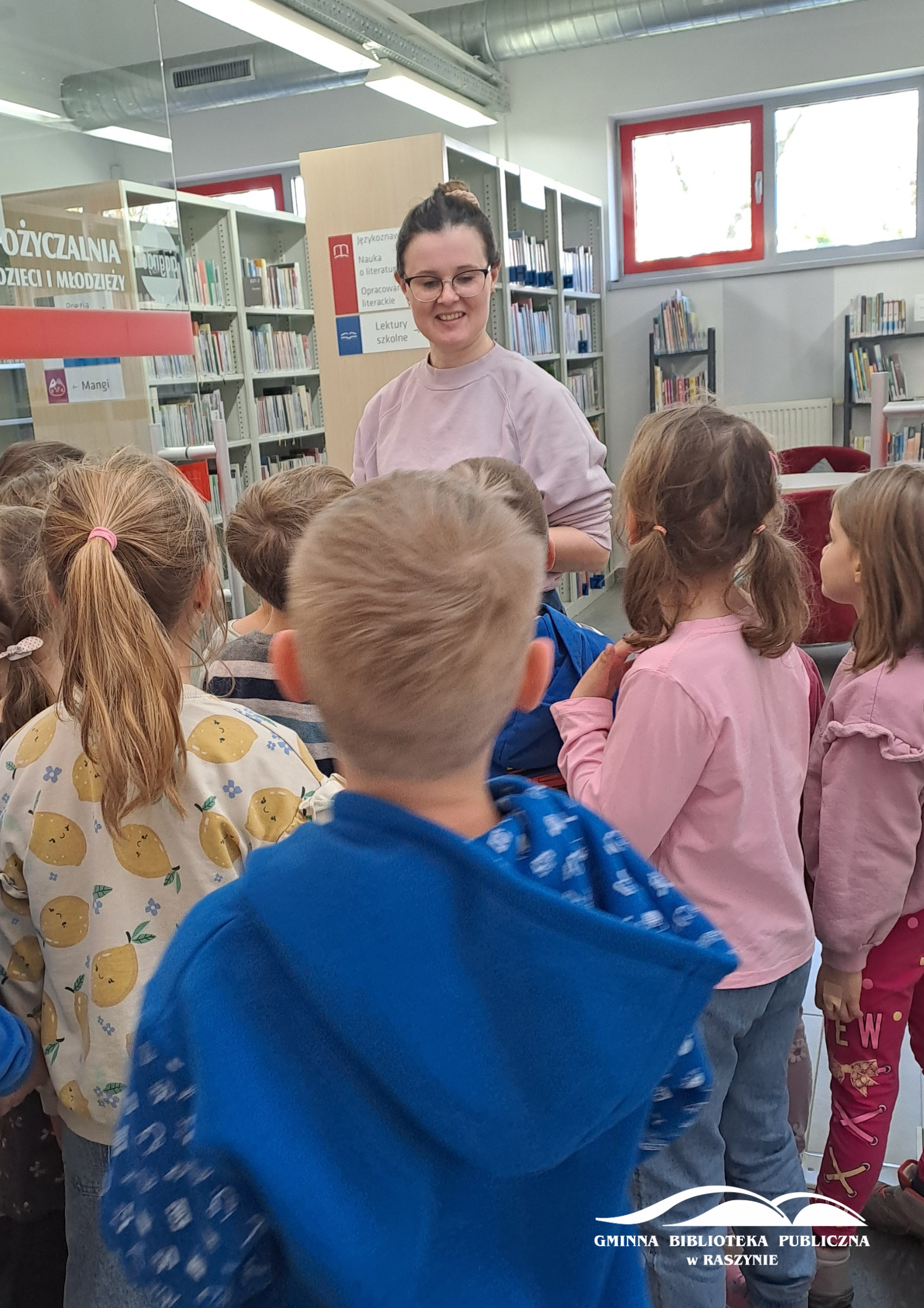 Dzieci zwiedzają bibliotekę - widoczna bibliotekarka na tle działu dla najmłodszych