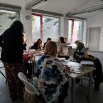 Uczestnicy warsztatów kolażowych wraz z prowadzącą siedzą przy stolikach z materiałami