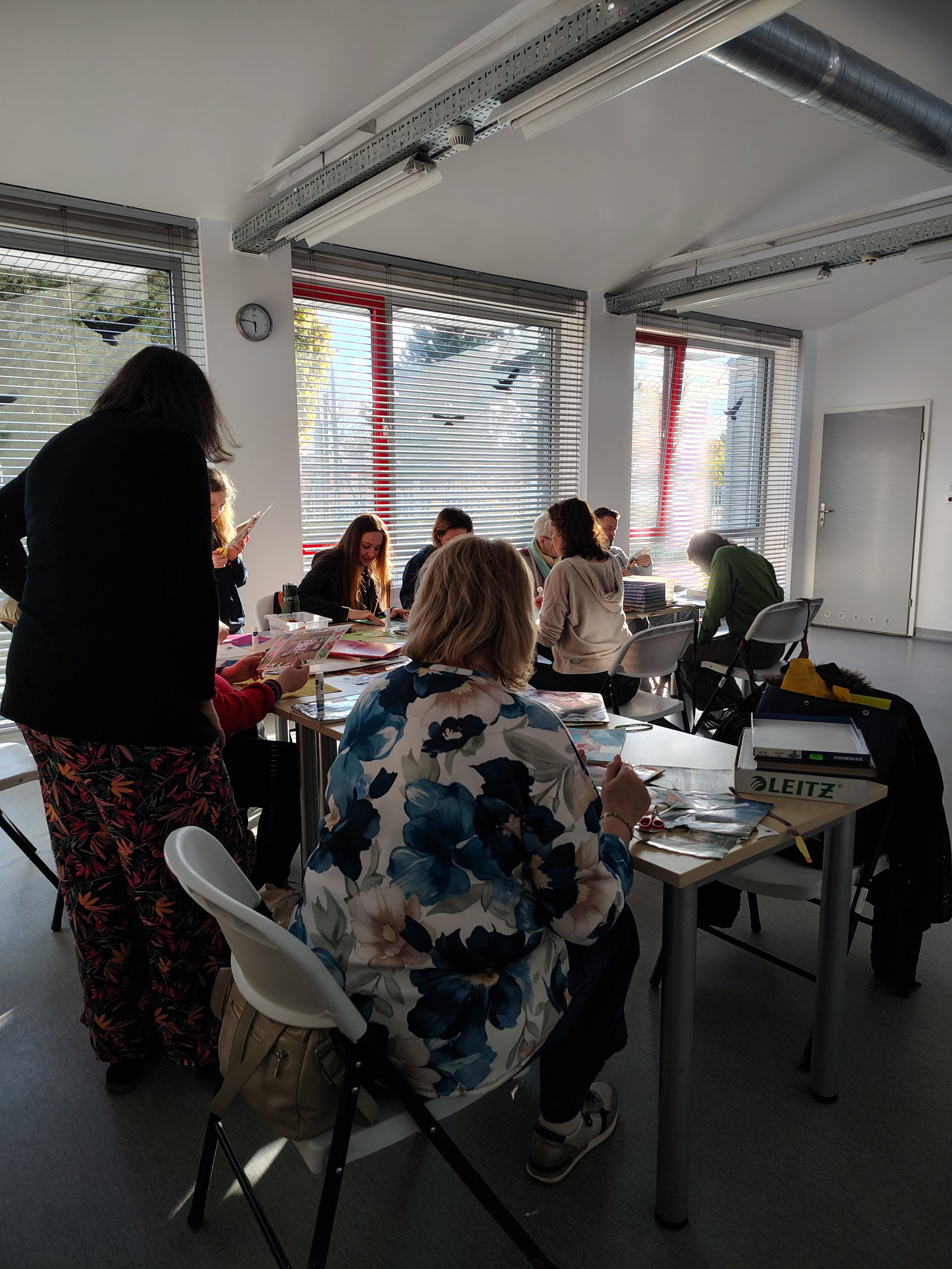 Uczestnicy warsztatów kolażowych wraz z prowadzącą siedzą przy stolikach z materiałami