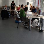 Grupa uczestników warsztatów kolażowych wraz z prowadzącą siedzą przy stolikach z materiałami