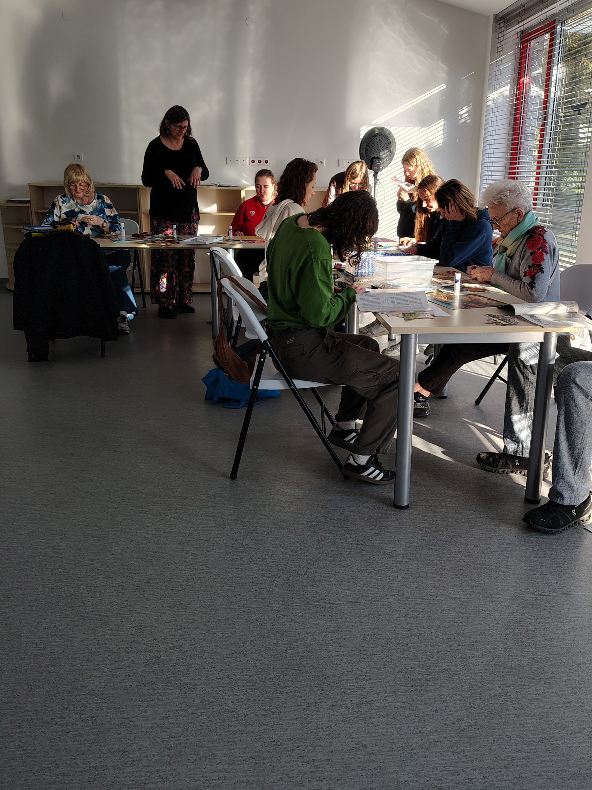 Grupa uczestników warsztatów kolażowych wraz z prowadzącą siedzą przy stolikach z materiałami