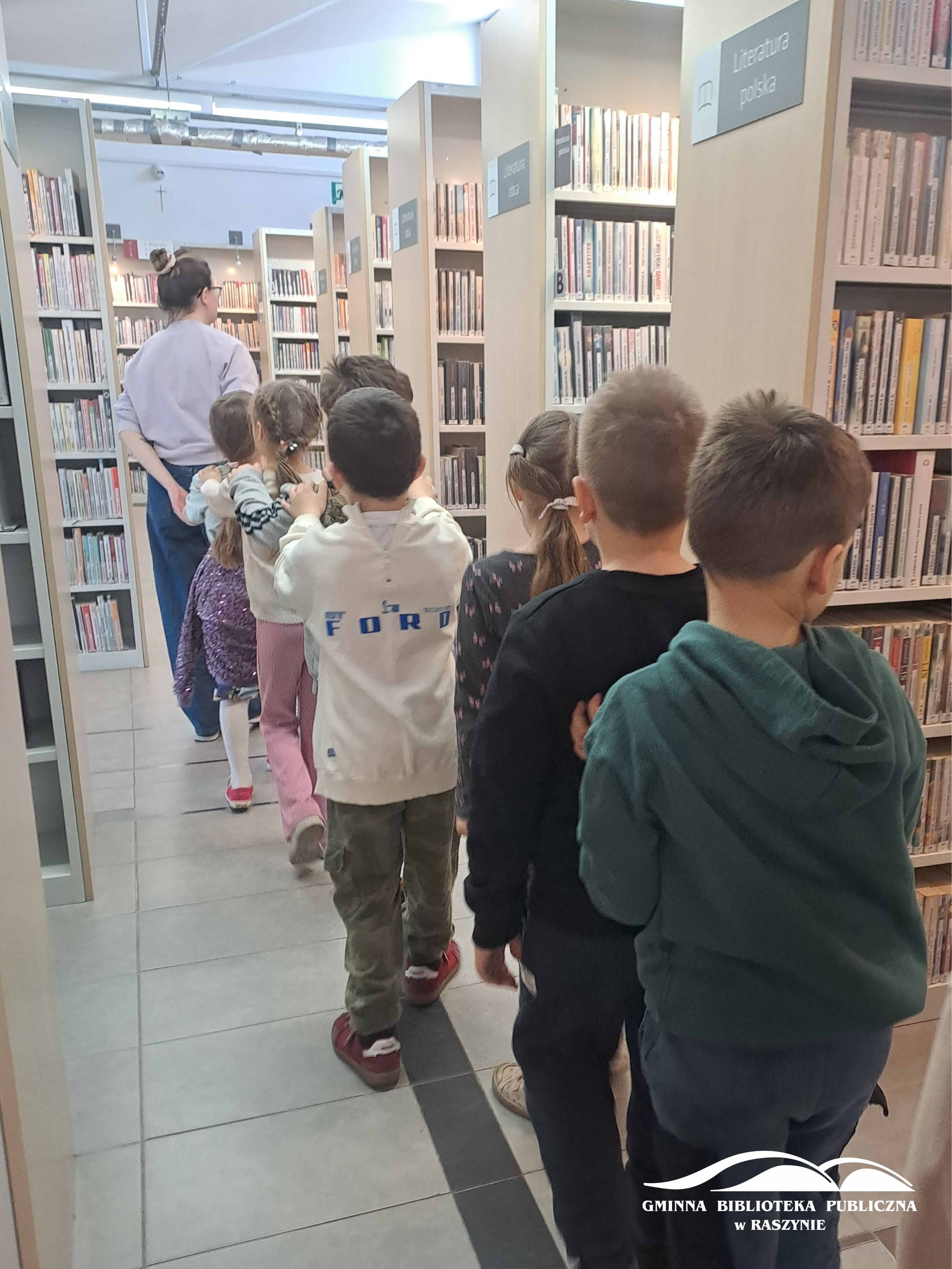 Dzieci podczas zwiedzania biblioteki - idą w rzędzie między regałami