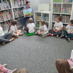 Dzieci siedzą w kole na dywanie, bibliotekarka pokazuje ilustrację z książki