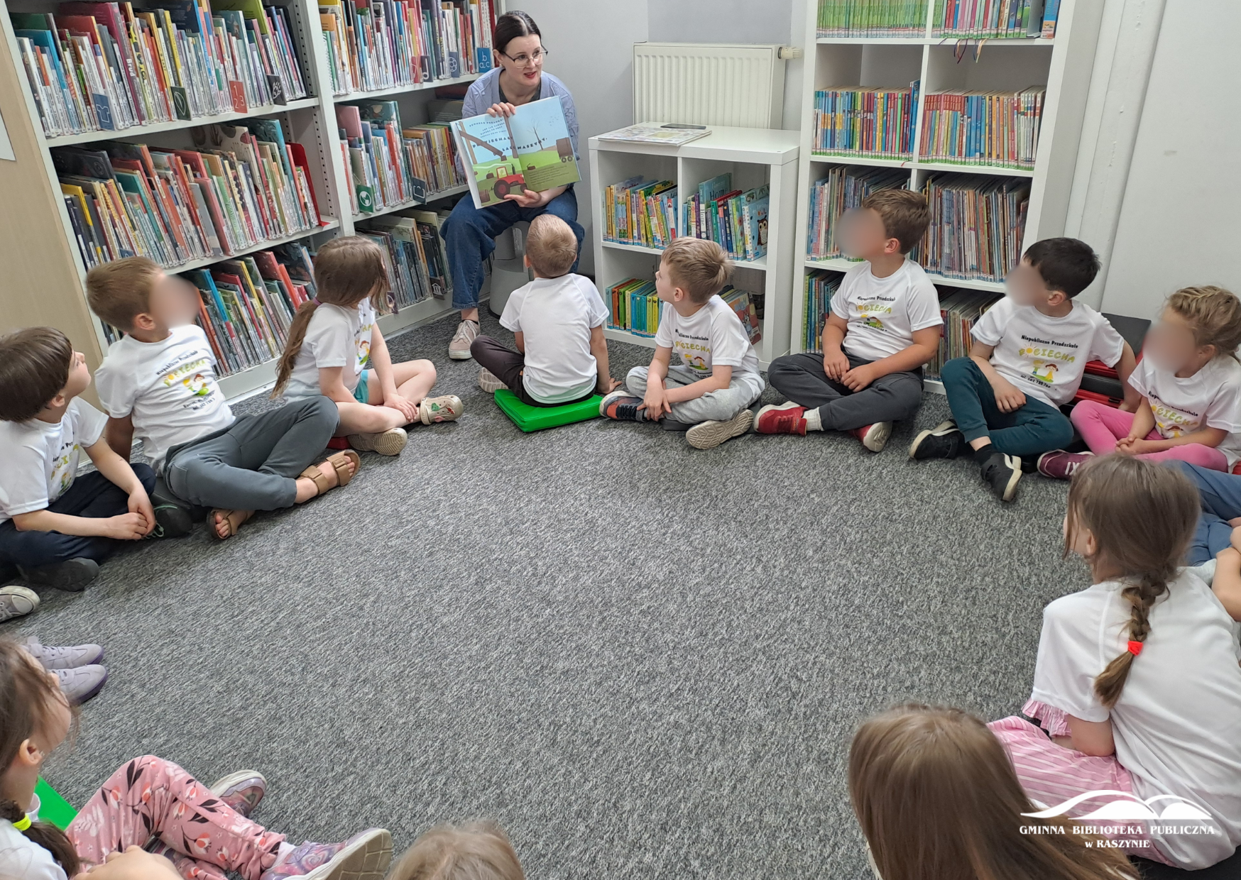 Dzieci siedzą w kole na dywanie, bibliotekarka pokazuje ilustrację z książki