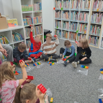 Dzieci siedzą w koło na dywanie, trzymają w uniesionych dłoniach czerwone klocki Lego