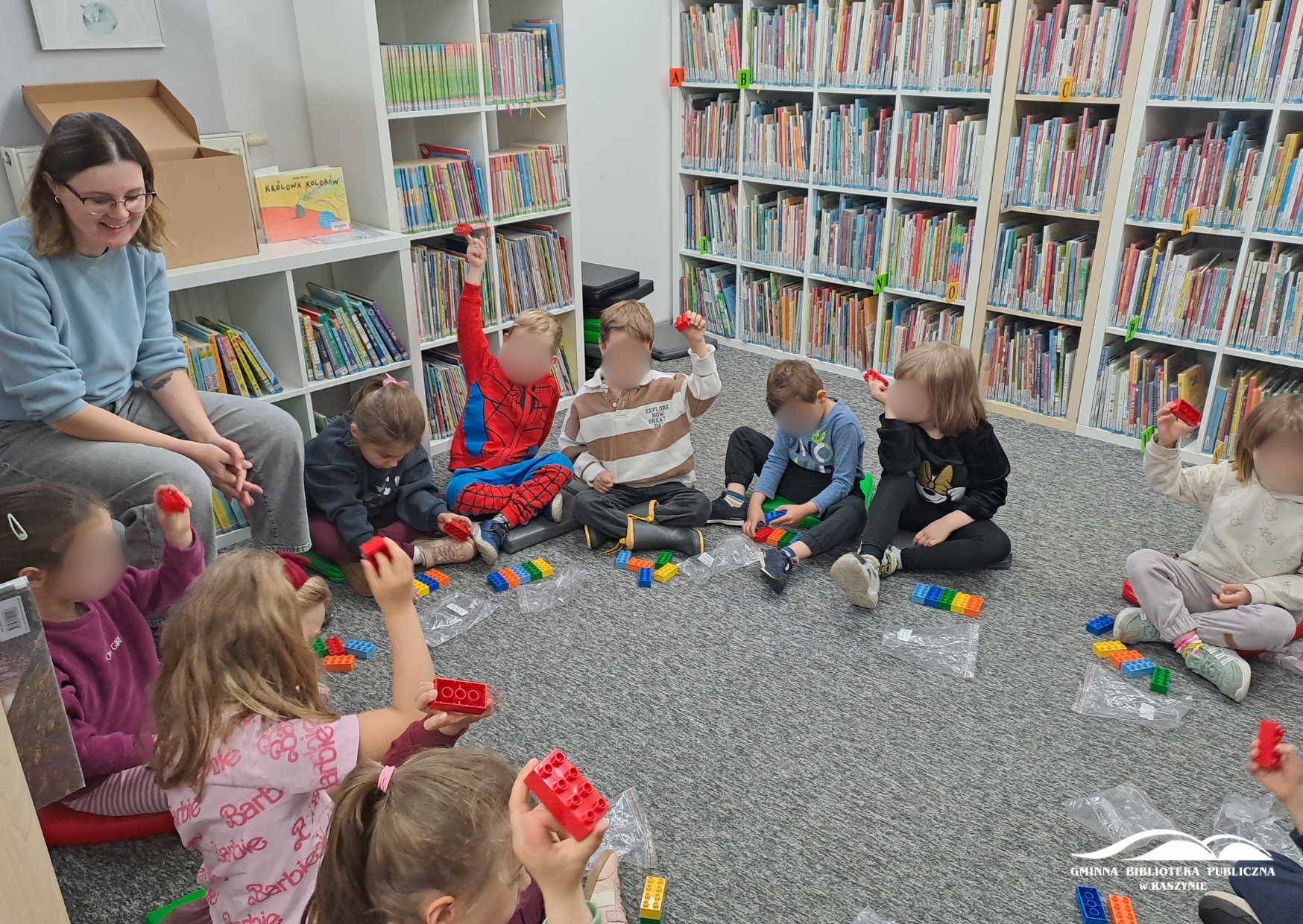 Dzieci siedzą w koło na dywanie, trzymają w uniesionych dłoniach czerwone klocki Lego