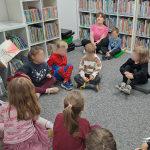 Dzieci siedzą w koło na dywanie, bibliotekarka pokazuje ilustrację z książki