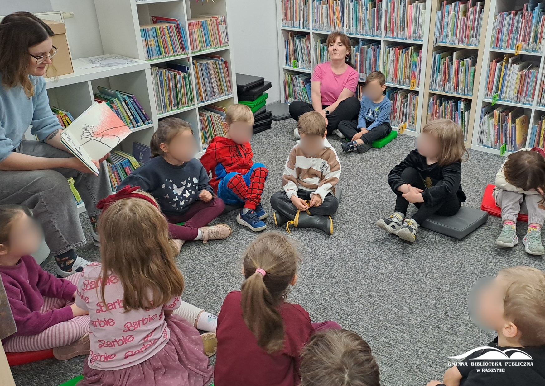 Dzieci siedzą w koło na dywanie, bibliotekarka pokazuje ilustrację z książki