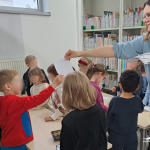 Dzieci stoją przy małych stolikach, bibliotekarka rozdaje karty pracy