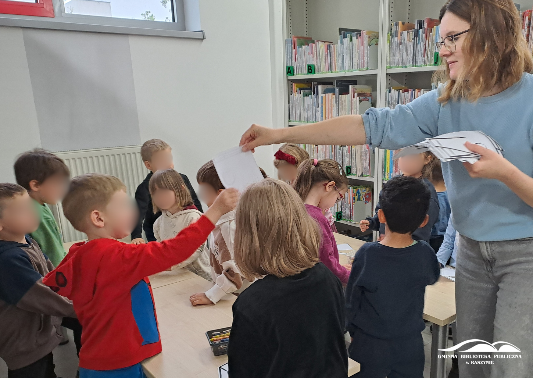 Dzieci stoją przy małych stolikach, bibliotekarka rozdaje karty pracy