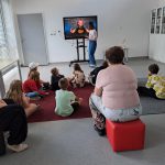 Grupa dzieci wraz z wychowawczyniami siedzi przed ekranem z filmikiem o historii komiksu