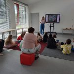 Bibliotekarka stoi przy ekranie, dzieci siedzą na dywanie
