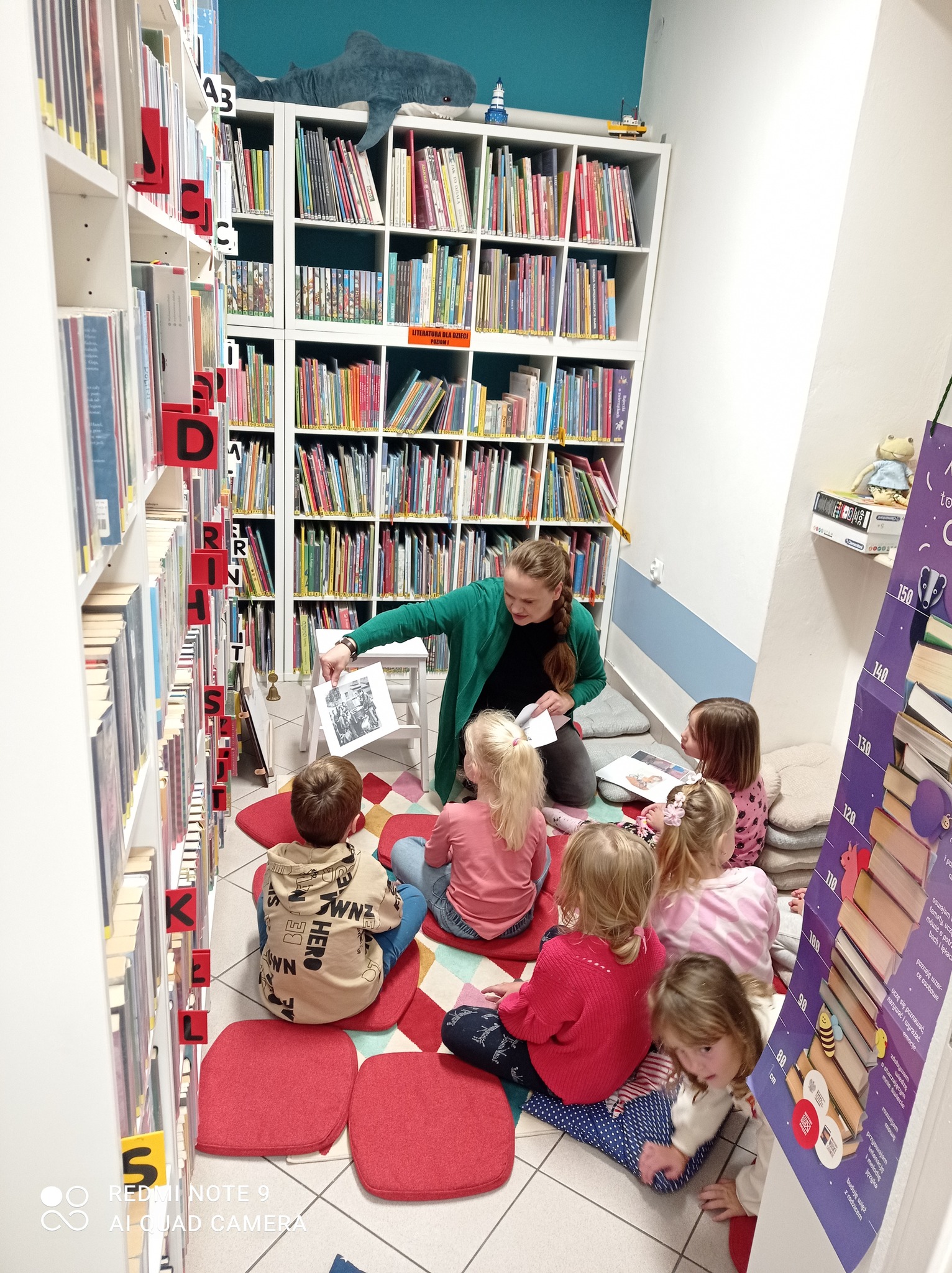 Bibliotekarka pokazuje kolejną planszę w teatrzyku kamishibai, przed nią siedzi grupa zasłuchanych dzieci