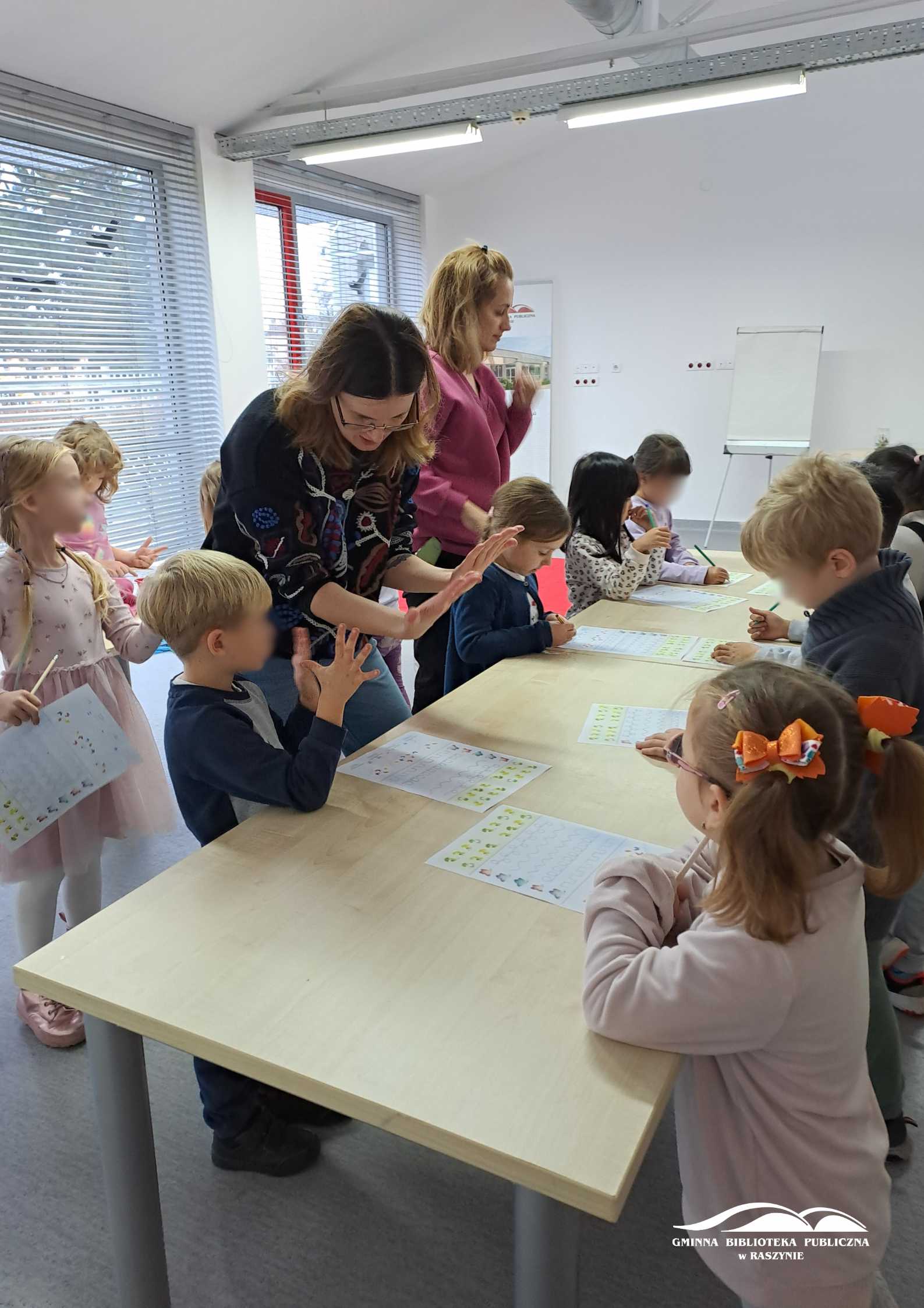 Grupa dzieci stoi z kartkami z zadaniami wokół stołu, bibliotekarka pokazuje, jak liczyć na palcach