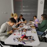 Grupa kobiet siedzi przy stole z rozłożonymi wycinankami, gazetami i papierem kolorowym