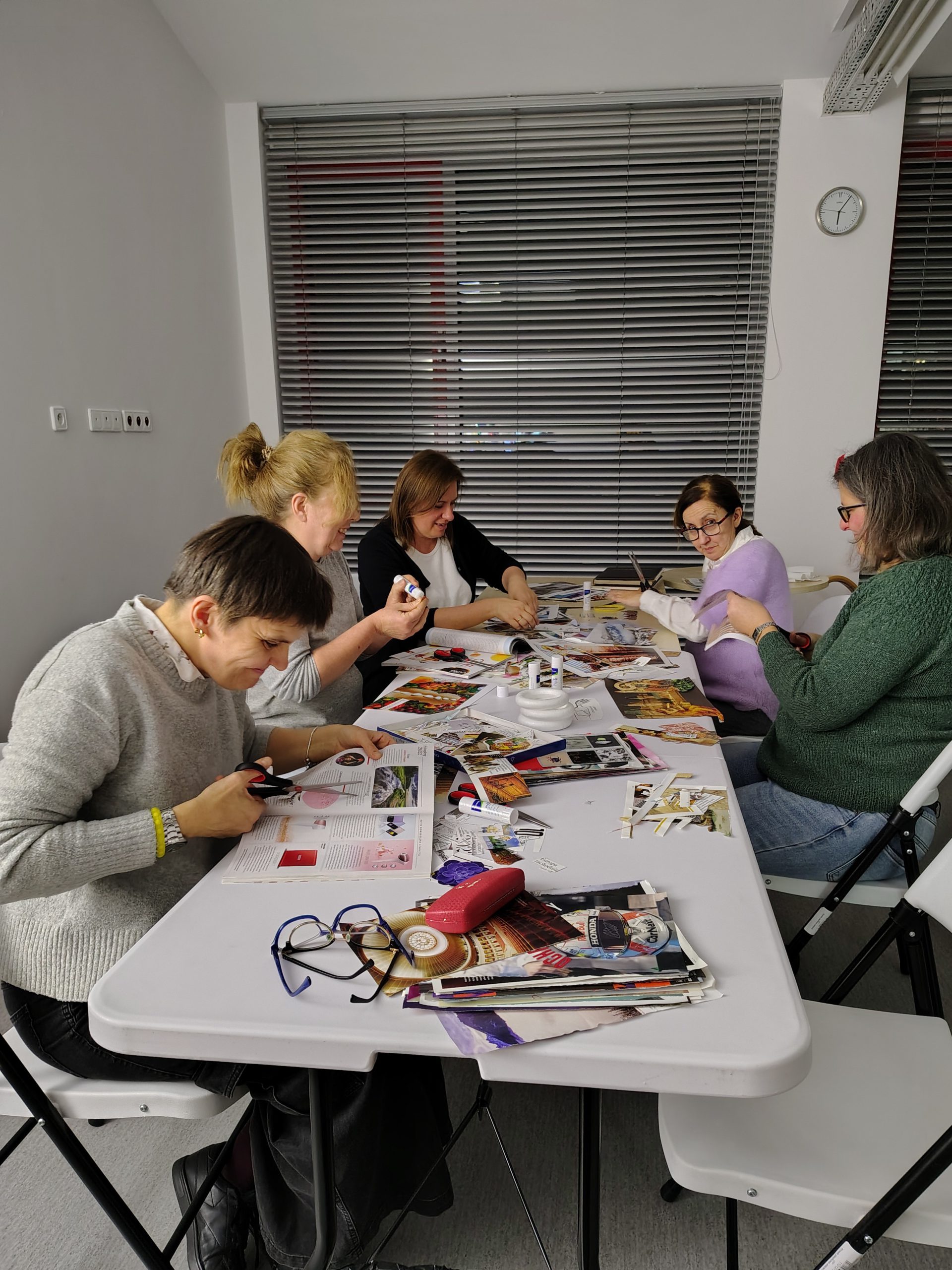 Grupa kobiet siedzi przy stole z rozłożonymi wycinankami, gazetami i papierem kolorowym