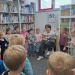 Grupa dzieci stoi w kąciku dla najmłodszych, bibliotekarka śpiewa i gra na ukulele