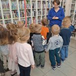 Grupka przedszkolaków wraz z bibliotekarką stoi przy dziale z audiobookami