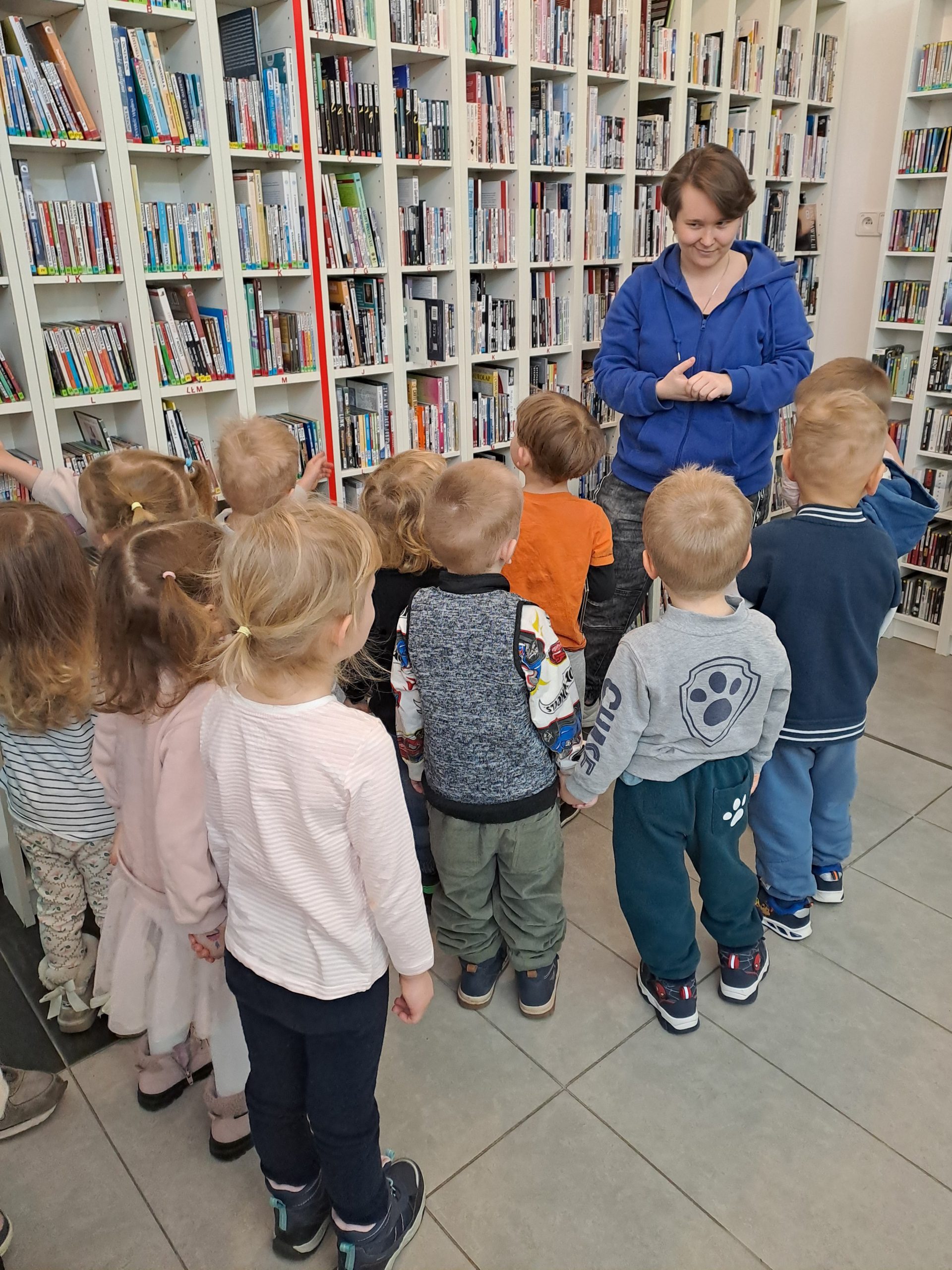 Grupka przedszkolaków wraz z bibliotekarką stoi przy dziale z audiobookami