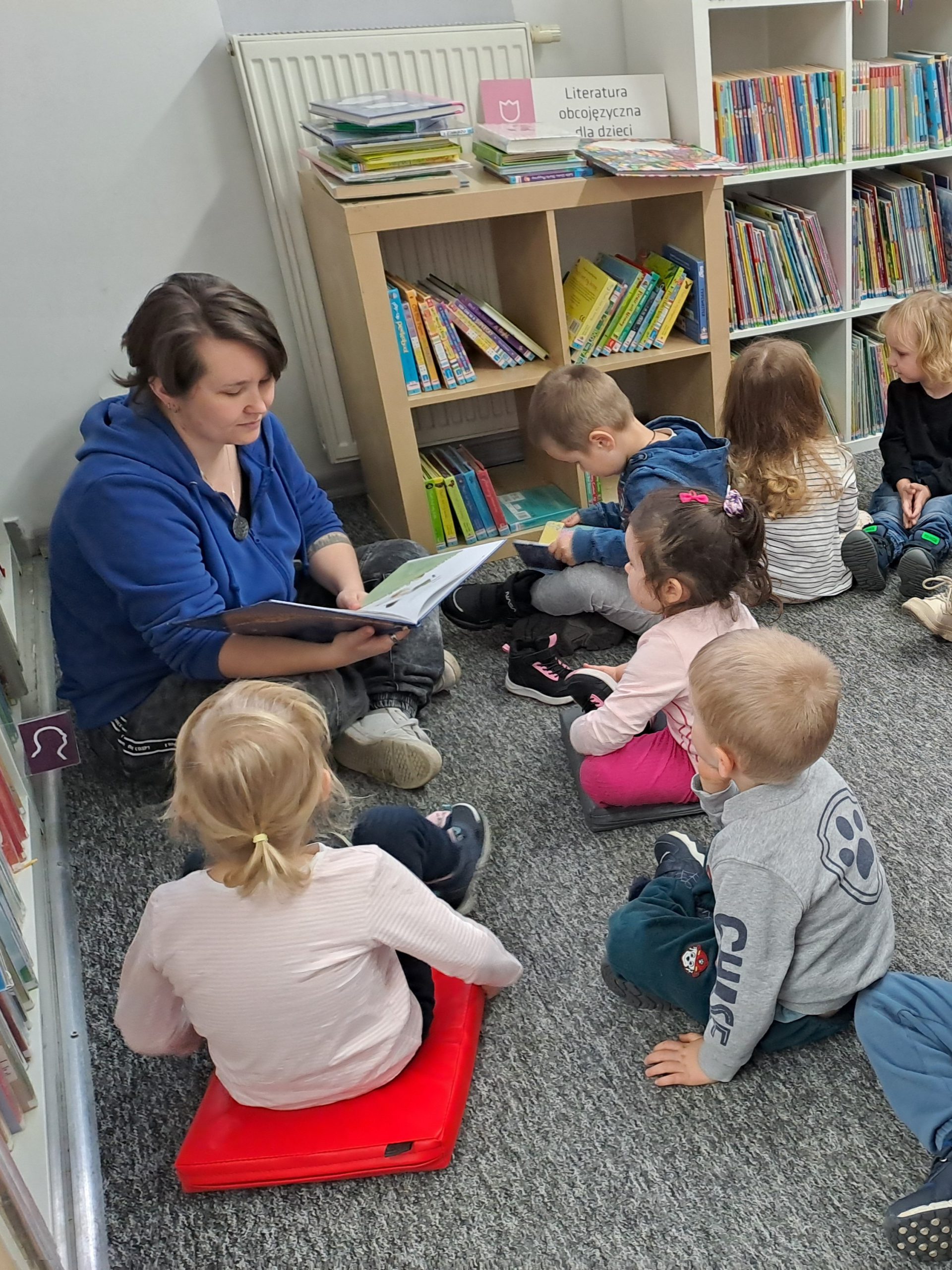 Bibliotekarka czyta książeczkę, grupka dzieci siedzi przed nią na dywanie