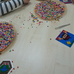 Zbliżenie na stół - widać powstające prace i dwie miseczki z koralikami